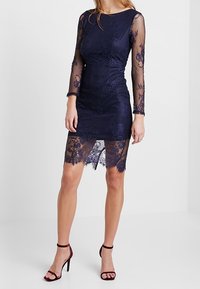 Mörkblå spetsklänning med långa genomskinliga ärmar, figursydd överdel och vågformad spetskant. Har intrikata blommönster och en V-formad halsringning.