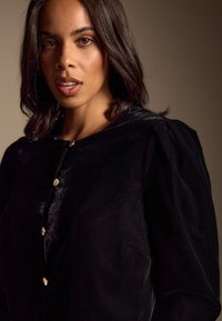 Zwarte fluwelen blouse met een ronde halslijn, knopen aan de voorkant en licht opgeblazen mouwen, voorzien van gouden knopen en een gladde textuur.