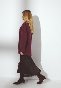 Blazer bordeaux sur une jupe noire transparente, associé à des bottes à talons marron. La tenue comporte des manches longues et une silhouette fluide.