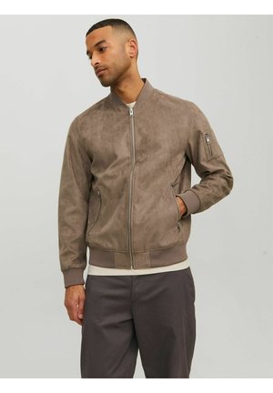 Mann trägt taupefarbene Wildleder-Bomberjacke mit Reißverschlusstaschen und dunkler Hose, steht vor schlichtem hellem Hintergrund.