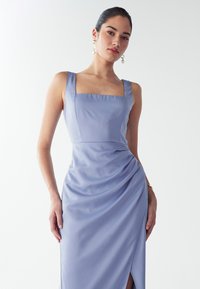 Kobieta ubrana w bezrękawnikową lawendową sukienkę z kwadratowym dekoltem i bocznym marszczeniem, uzupełniona długimi perłowymi kolczykami i bransoletką.