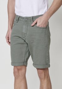 Shorts en denim vert olive avec une coupe ajustée, ourlets roulés et passants de ceinture, présentant des poches avant et une texture subtile.