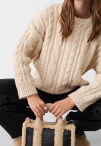 Kermainen, palmikkoneulottu villapaita, jossa on resorit hihansuissa, yhdistetty mustiin farkkuihin. Beige pehmeäpintainen kangaskassi, jossa mustat nahkaiset yksityiskohdat.