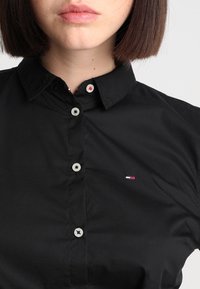 Černá košile se zapínáním na knoflíky a límečkem, s světle zbarvenými knoflíky a malým logem v červené, bílé a modré na levé straně. Hladká tkanina.