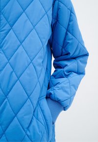 La chaqueta de plumas acolchada azul presenta un patrón de diamantes, puños acanalados y bolsillos laterales. El material parece suave y ligero.