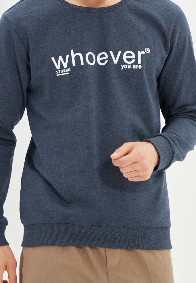 Personne portant un sweat-shirt bleu marine avec le texte « qui que vous soyez » en blanc sur la poitrine, associé à un pantalon marron clair.