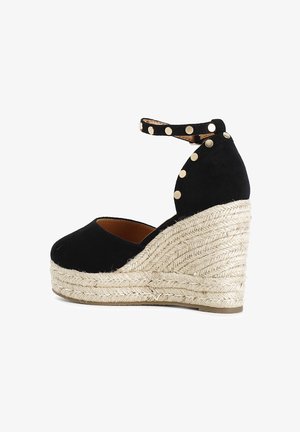 Espadrille negra de ante con cuña, suela de cuerda de yute, tira en el tobillo con tachuelas doradas, puntera redondeada y detalles texturizados a lo largo del costado.