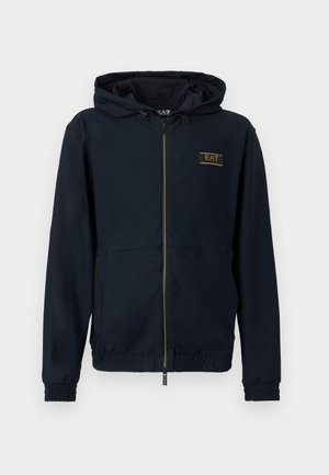 Chaqueta azul marino con capucha y cierre de cremallera, tela texturizada, puños elásticos y banda en la cintura, con un logo dorado en el pecho izquierdo.