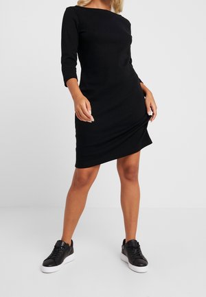 Czarna sukienka z długim rękawem, okrągłym dekoltem, dopasowanym krojem i lekko zaokrąglonym dołem. Zestawiona z czarnymi trampkami na jednolitym tle.