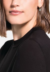 Kvinna med kort brunt hår som bär en svart topp och guldörhängen i tårform med blå ädelstenar, med slutna läppar och neutral uttryck.