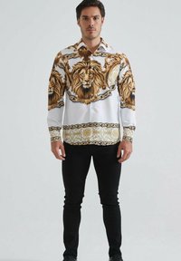 Homme debout portant une chemise blanche à boutons ornée de grands visages de lion détaillés et de motifs en chaîne dorée, associée à un pantalon noir et des chaussures.