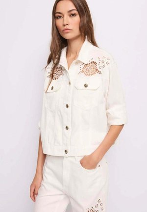 Donna che indossa una giacca bianca con bottoni e ricamo floreale traforato sulle spalle, abbinata a pantaloni bianchi con la mano in tasca.