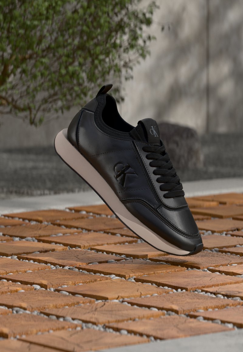Baskets en cuir noir avec semelle blanche, logo CK sur le côté et sur la languette, flottant au-dessus d'un sol en carreaux de bois à l'extérieur avec un feuillage vert en arrière-plan.