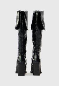 Pair of black high-heeled leather knee boots with fold-over cuffs and side zippers, shown from the back on a white background.