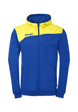 Veste à fermeture éclair bleue avec motif géométrique jaune sur les épaules et la capuche, avec le logo Kempa sur la poitrine gauche.
