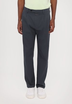 Person, die dunkelgraue, gerade geschnittene Hose, weiße Sneakers und ein helles, in die Hose gestecktes Hemd trägt, steht vor einem schlichten Hintergrund.