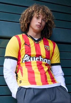 Red and yellow striped soccer jersey with a black collar, featuring a logo and text. Worn over a white long-sleeve shirt, with gray pants.