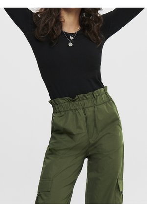 Femme levant les bras portant un haut noir à manches longues, des colliers argentés superposés et un pantalon cargo vert olive taille haute avec une ceinture froncée.