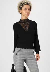 Black sweater with lace high neck, scalloped hem, and long sleeves, paired with gray checkered pants featuring zippered pockets.