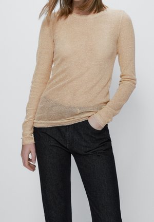 Femme portant un haut beige texturé à manches longues et un pantalon noir, debout avec une main dans la poche devant un fond uni.