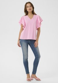 Roza majica s V-izrezom in volančastimi kratkimi rokavi, v kombinaciji z modrimi oprijetimi kavbojkami in roza sandali s peto, na enobarvni beli ozadju.