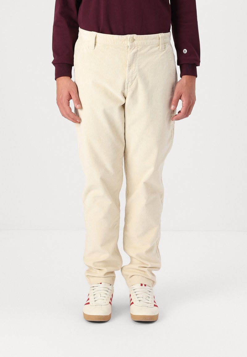 Pantaloni in velluto a coste beige con vestibilità slim, dotati di taschini anteriori e chiusura con bottone, abbinati a sneakers bianche con strisce rosse.