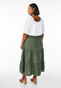 Haut en eyelet blanc avec des manches bouffantes associé à une jupe verte olive à volants. Des sandales à plateforme marron complètent la tenue.