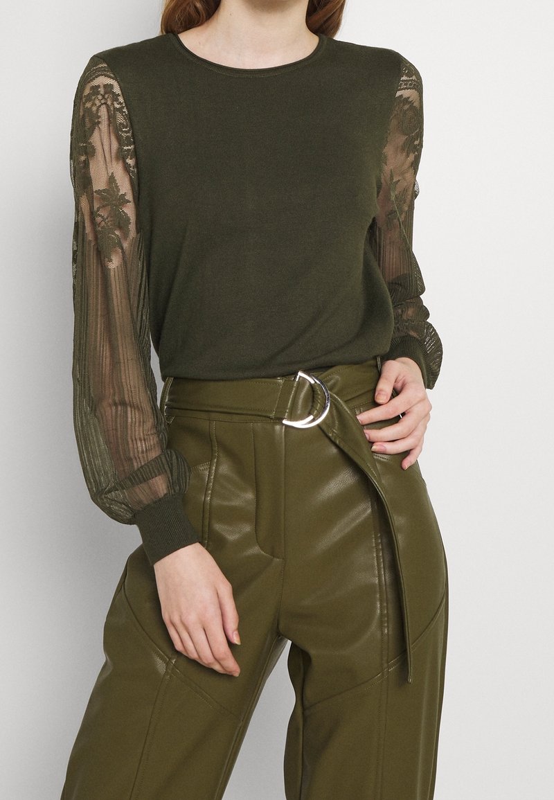 Femme portant un chemisier vert olive avec des manches en dentelle transparente et un pantalon en cuir vert olive taille haute avec une ceinture assortie.