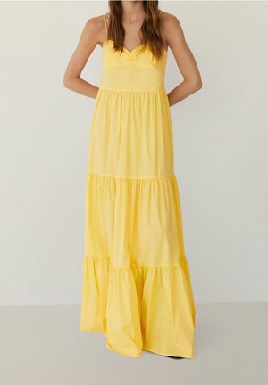 Frau trägt ein ärmelloses, gestuftes gelbes Maxikleid mit tailliertem Oberteil und fließendem Rock, steht mit den Händen hinter dem Rücken.