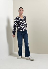Blouse à motif géométrique bleu marine et blanc, coupe décontractée, boutonnière devant ; associée à un jean droit bleu et des baskets blanches avec des accents.