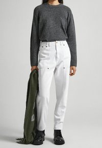 Maglione grigio a maglia, pantaloni bianchi a vita alta con tasche frontali e stivali neri stringati. Tiene una sciarpa verde con scritte bianche.