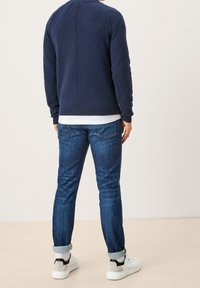 Homme portant un pull bleu foncé, un t-shirt blanc, un jean bleu retroussé, des chaussettes blanches et des baskets blanches, debout de dos.