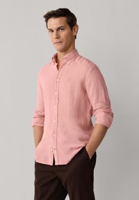 Homme portant une chemise à boutons rose clair aux manches retroussées et un pantalon marron foncé, debout les mains dans les poches devant un fond uni.
