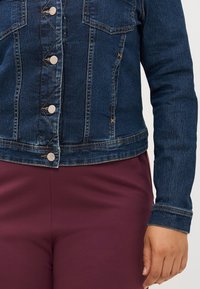 Denim jakna s gumbi, oblečena čez bordo hlače. Poudarek na predelu prsi prikazuje podrobnosti tkanine in šivov.