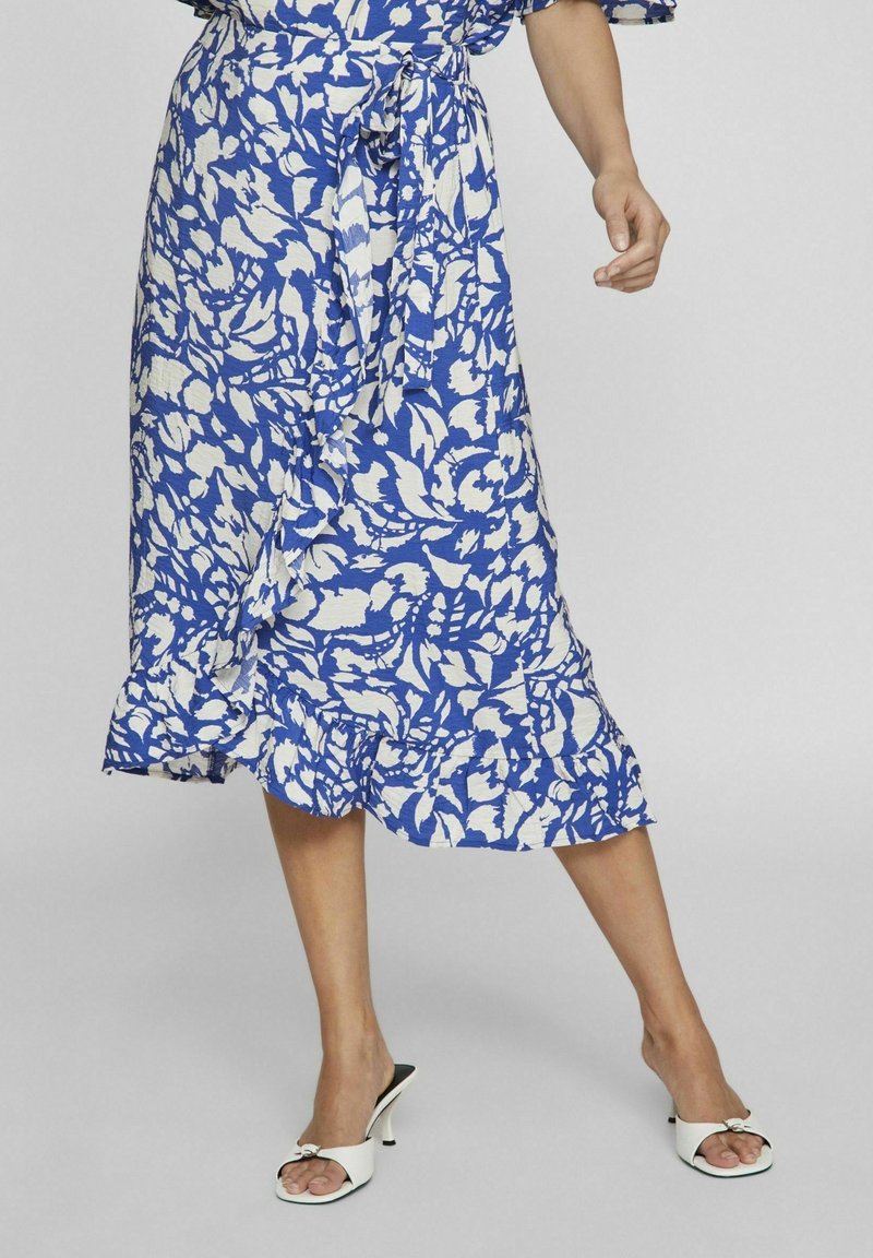 Modrá a bílá sukně s květinovým vzorem, texturovaná látka, volánkový lem, zavázaný pas, v kombinaci s bílými sandály na otevřené špičce.