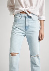 Woman wearing light blue ripped jeans and a pink long-sleeve shirt tied at the waist, standing with one hand in pocket.