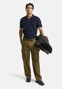 Polo shirt marine, pantalon cargo vert olive avec poches latérales, ceinture marron et une veste sombre, porté avec des chaussures noires.