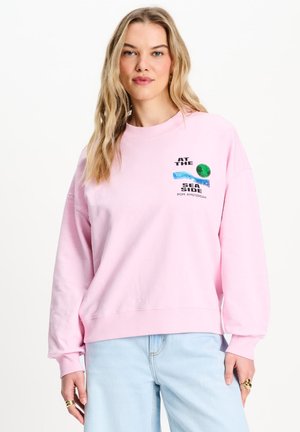 Jonge vrouw met lang blond haar, die een lichtroze sweatshirt met een kustafbeelding en lichtblauwe spijkerbroek draagt, staand tegen een witte achtergrond.