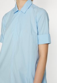 Camisa azul clara de mangas curtas com colarinho pontudo, textura suave e costura visível; apresenta um bolso lateral discreto e punhos enrolados.