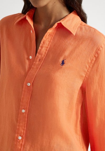 Sieviete valkā spilgti oranžu krekla kreklu ar zilas izšūta polo spēlētāja logo kreisajā krūtīs.