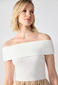 Bijela off-shoulder majica s rebrastom teksturom, glatkim materijalom i širokim trakama, u kombinaciji s bež suknjom s nabranim detaljima.