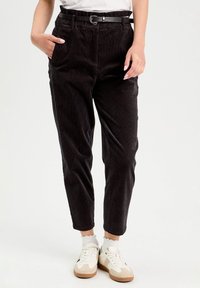 Pantalon en velours côtelé noir avec une coupe ajustée, doté d'une ceinture noire, de poches latérales et d'une texture douce. Associé à des baskets blanches.