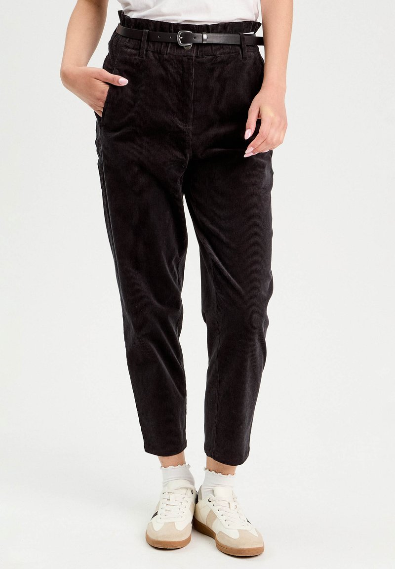 Pantalon en velours côtelé noir avec une coupe ajustée, doté d'une ceinture noire, de poches latérales et d'une texture douce. Associé à des baskets blanches.