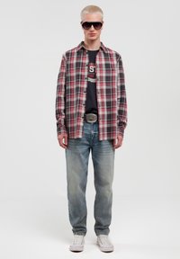 Rutet skjorte i rød, sort og hvid over en sort grafisk t-shirt, parret med lyseblå jeans og hvide sneakers. Store sorte solbriller og bælte.