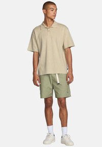 Beige kortærmet polo t-shirt med et subtilt mønster, parret med olivengrønne shorts med snøre og logo. Hvide sneakers fuldender looket.