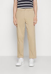 Chinos beige à coupe droite, avec passants pour ceinture, fermeture par bouton et poches latérales. Portés avec des baskets blanches.