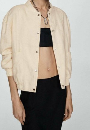 Personne portant une veste beige boutonnée sur un top bandeau noir et un pantalon noir, avec un collier orné d’un grand pendentif en argent.
