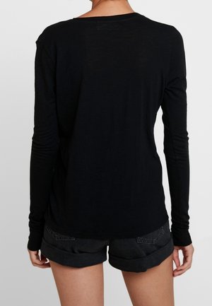 Henkilö pukeutuneena mustaan, vartalonmyötäiseen pitkähihnaiseen paitaan ja ylös käärityihin mustiin farkkushortseihin, nähtynä takaa yksiväristä taustaa vasten.