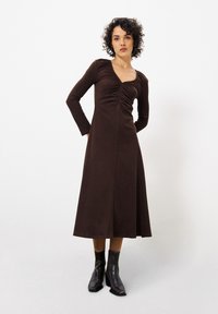 Braunes langärmeliges Kleid mit gerafftem Oberteil, A-Linien-Silhouette, glatter Textur und knielanger Länge, kombiniert mit schwarzen Knöchelstiefeln.