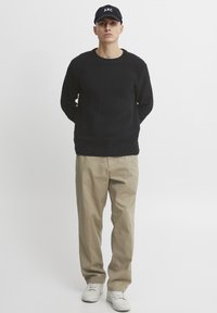 Black textured sweater, beige trousers, white sneakers, and a dark cap with "ARC" embroidered. Standing vertically against a plain background.